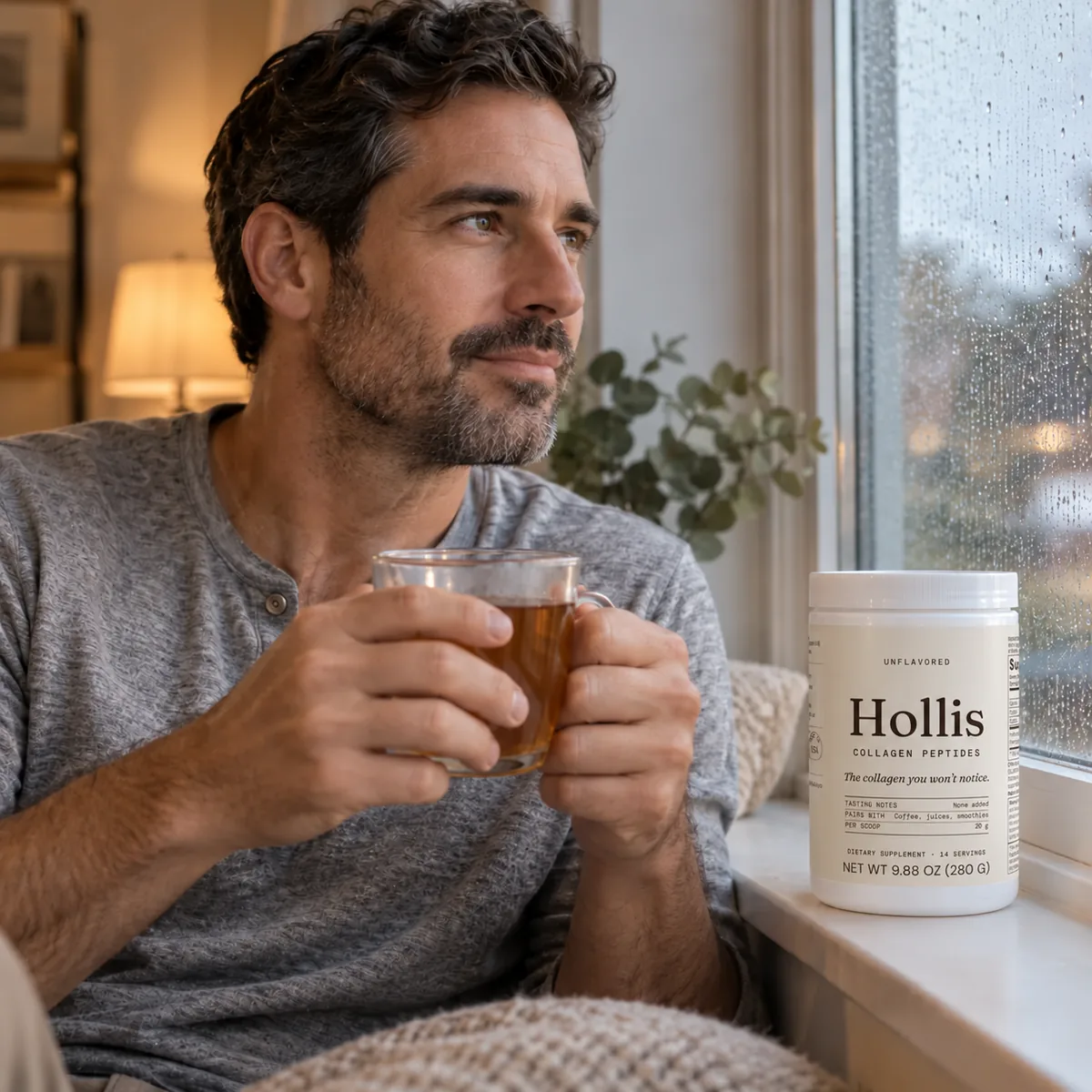 A man holding a cup of tea by a window, the Hollis container on the sill